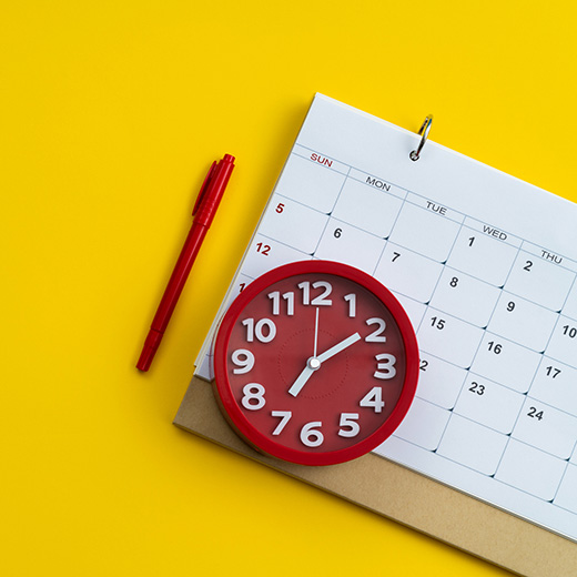 A red pen and red clock rest next to and on top of a white calendar over a bright yellow background.